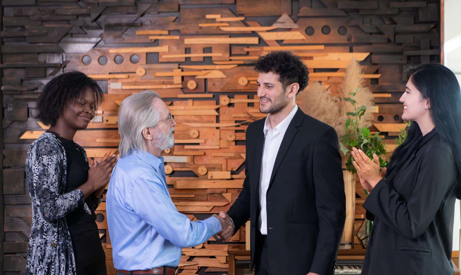 An older businessman shakes the hand of a young entrepreneur. Two colleagues applaud from the sidelines.