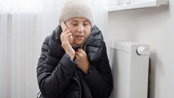 A woman in a woolly hat clutches her coat while sitting next to a radiator. She is on the phone calling for help with her broken boiler. 