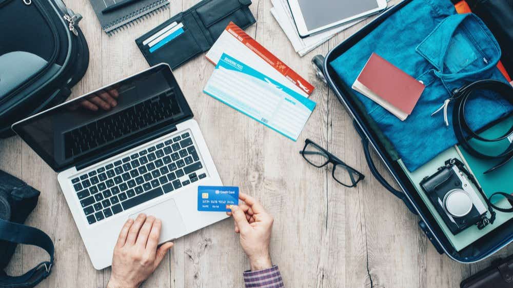 A man is at a laptop with a credit card and his suitcase is open next to him