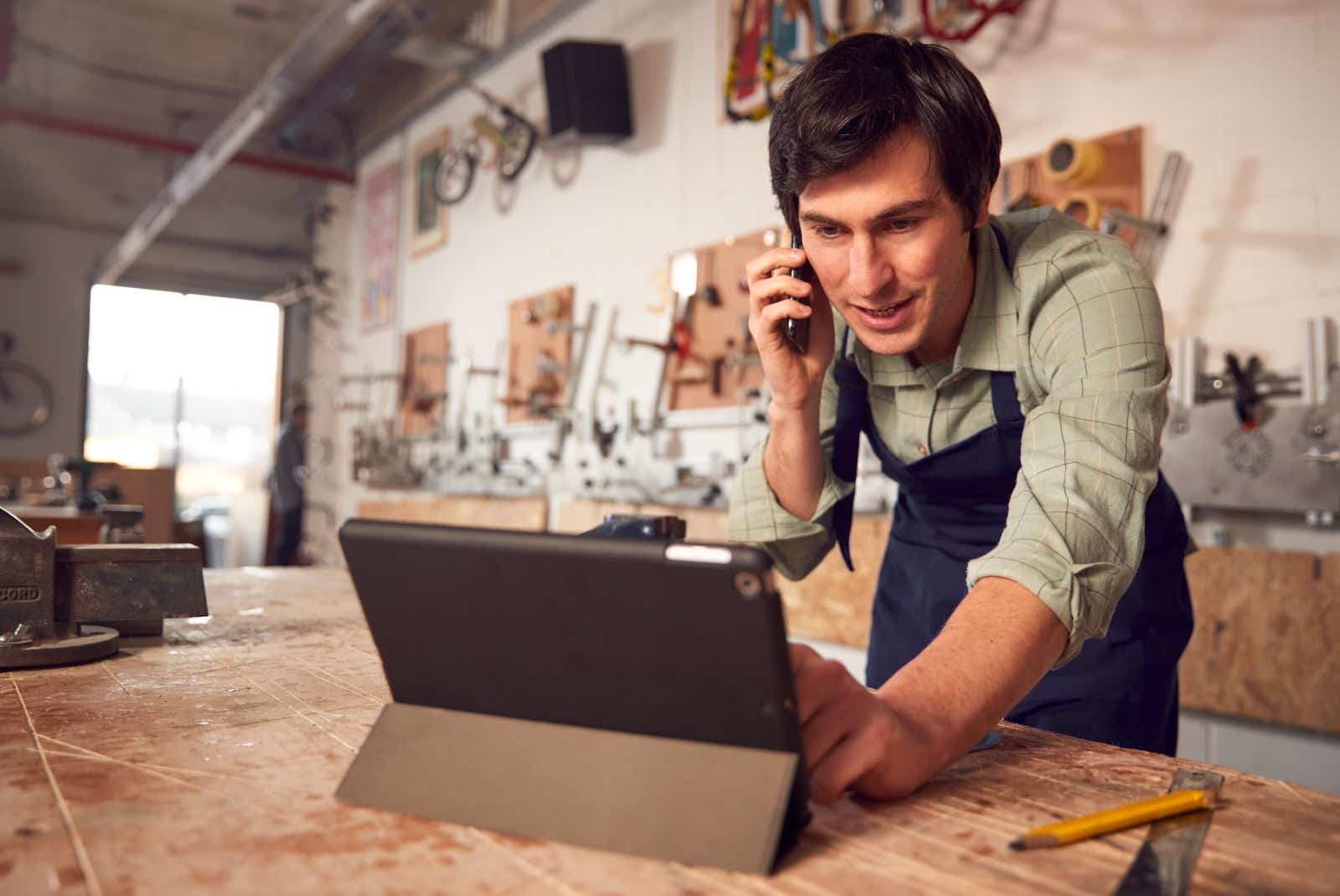 Business craftsman in a joinery shop using a laptop while on the phone. 