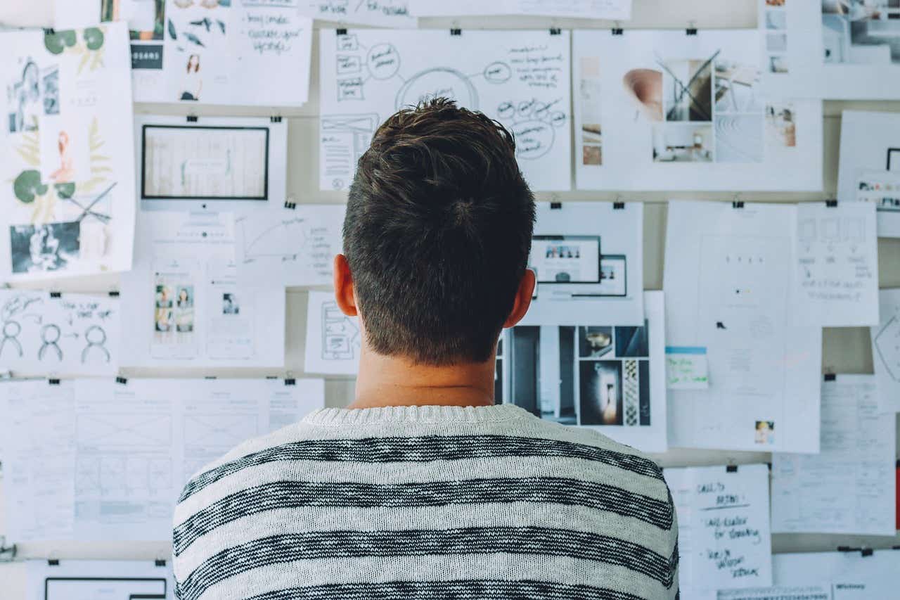 A man is facing a whiteboard with lots of writing and mind maps on it. 