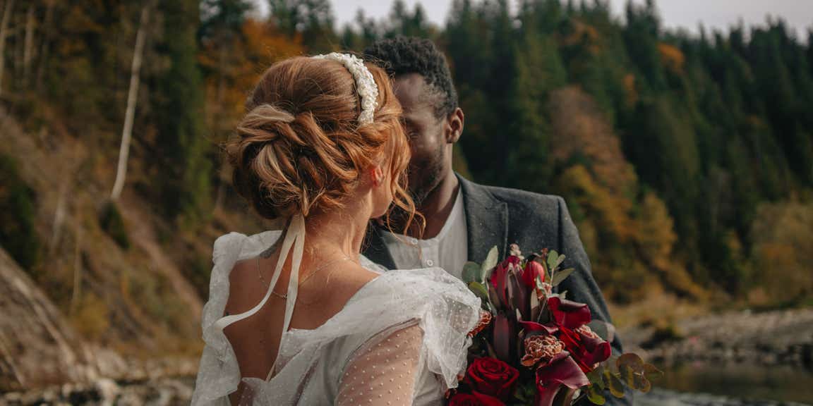 An interracial couple of newlyweds walks along the shore of a mountain river.