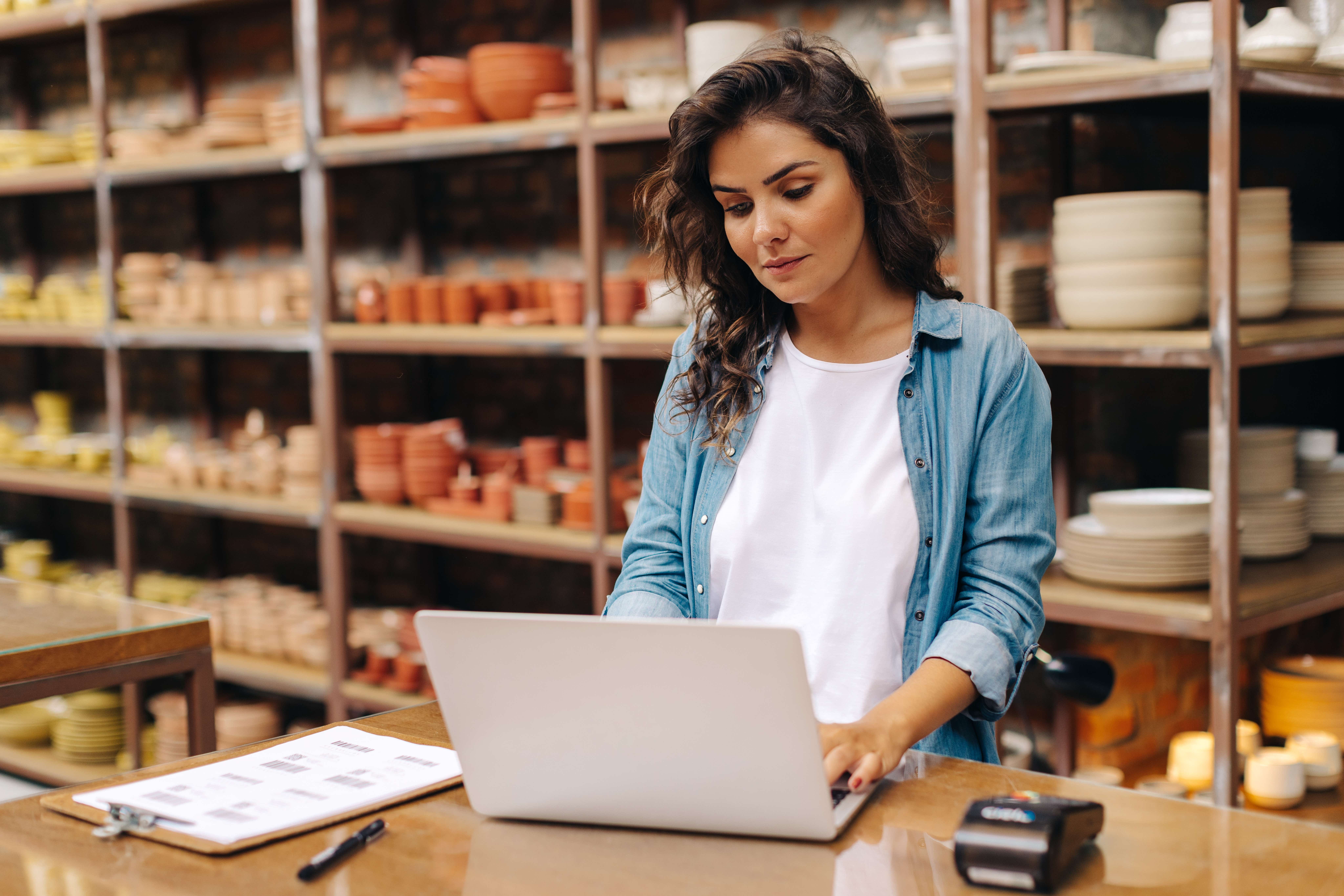 Self-employed woman using a laptop in a pottery shop, surrounded by shelves of ceramic items.