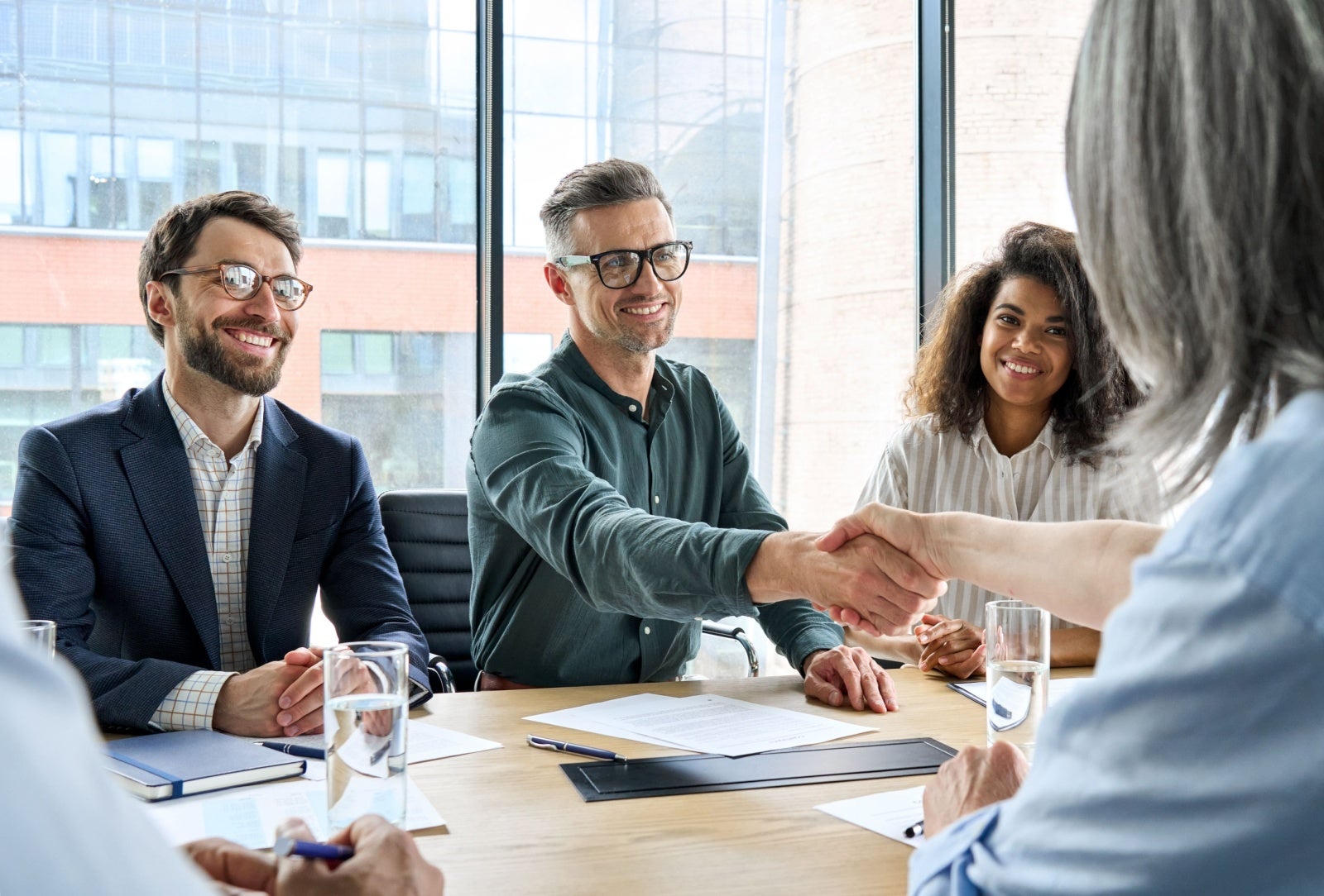 Smiling business people gathered around a conference table. In the middle, one man is shaking hands with the person opposite, signalling an agreement.
