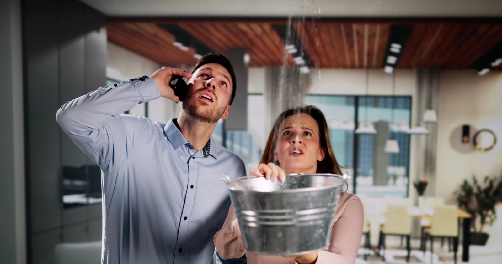 A couple phone for help as water pours in from their ceiling. The woman is trying to catch the water in a zinc bucket.