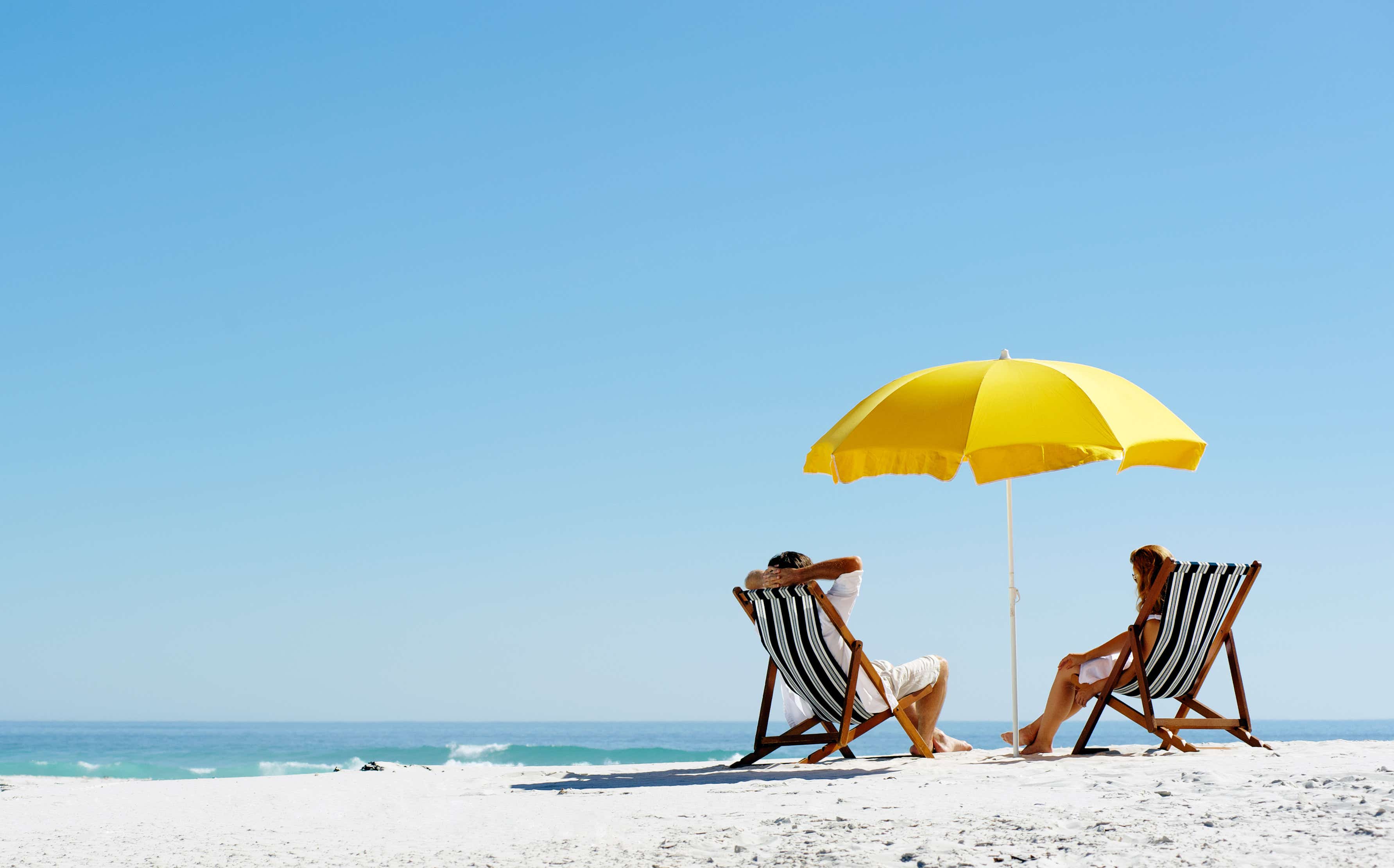 Couple on tropical island holiday relax in the sun on their deck chairs under a yellow umbrella.
