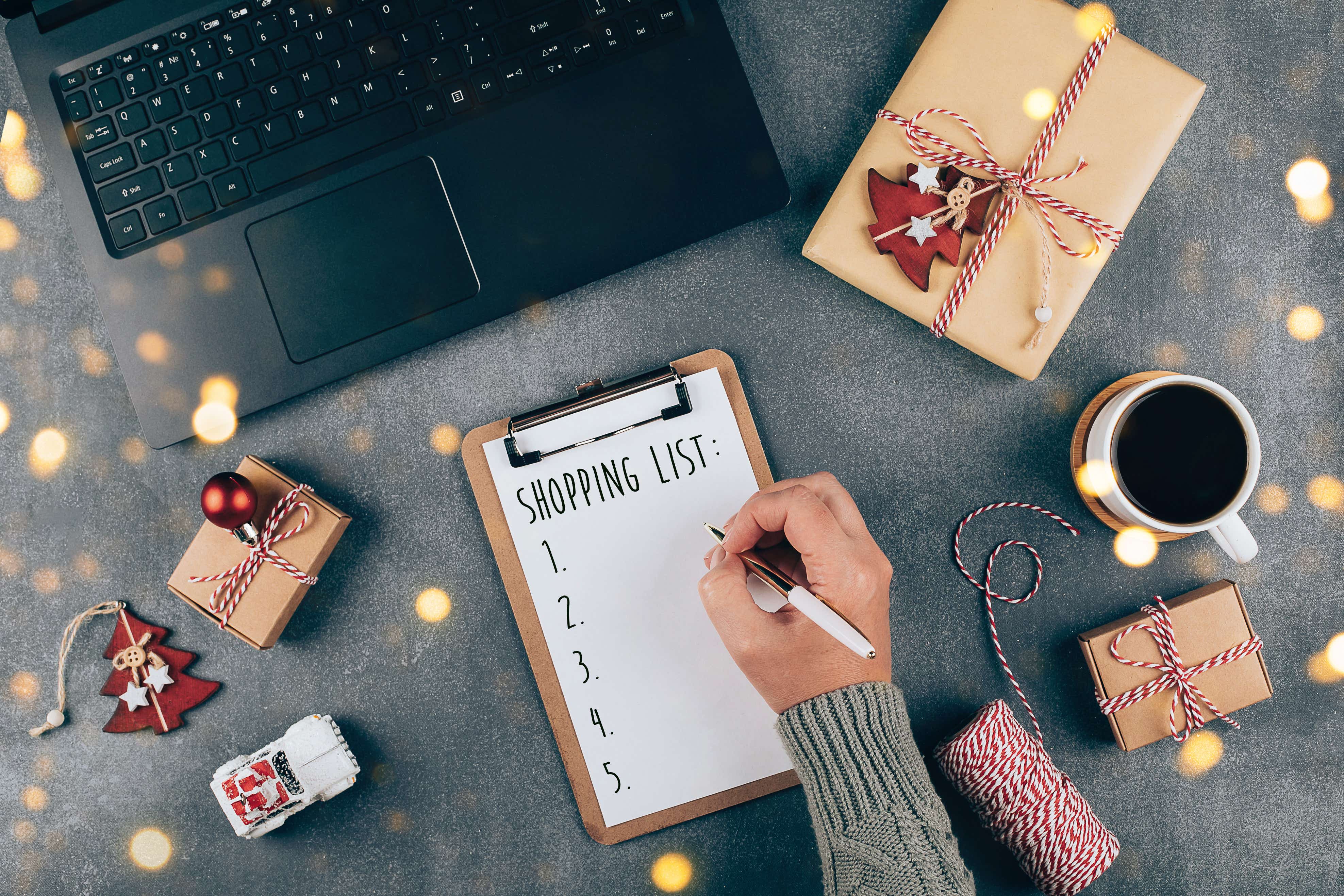 A laptop surrounded by Christmas decorations with someone writing a shopping list. 