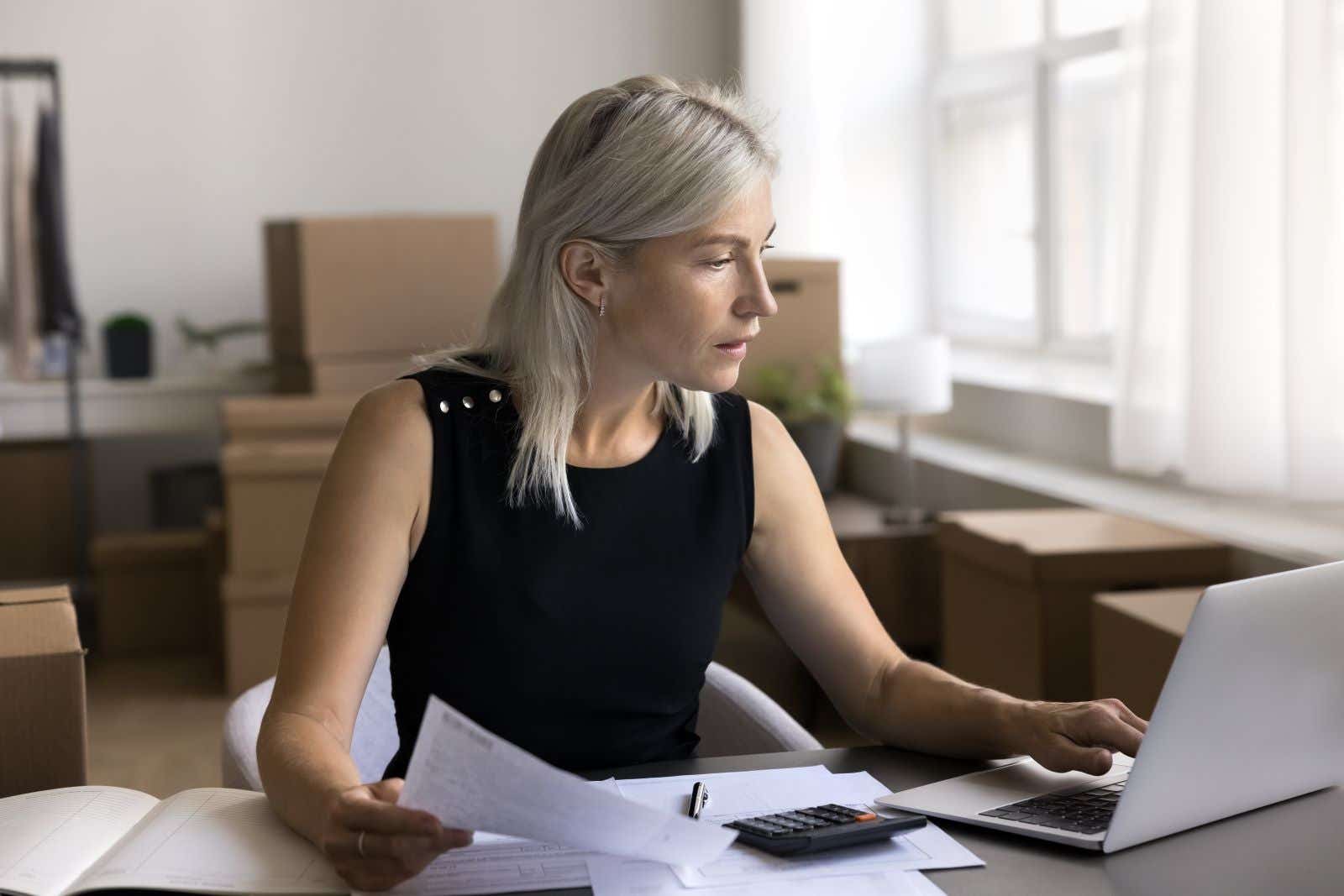 Woman using a laptop. She has paperwork in her hand and more on her desk.