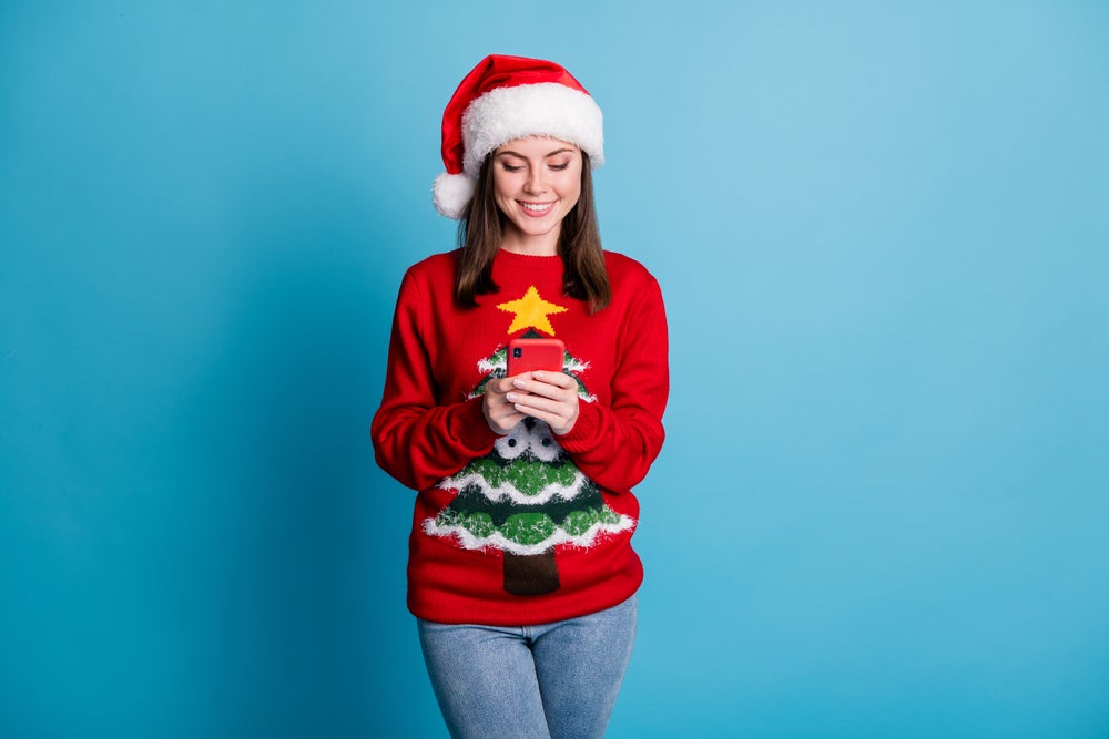 Woman is looking at her phone and is wearing a Christmas jumper and hat.