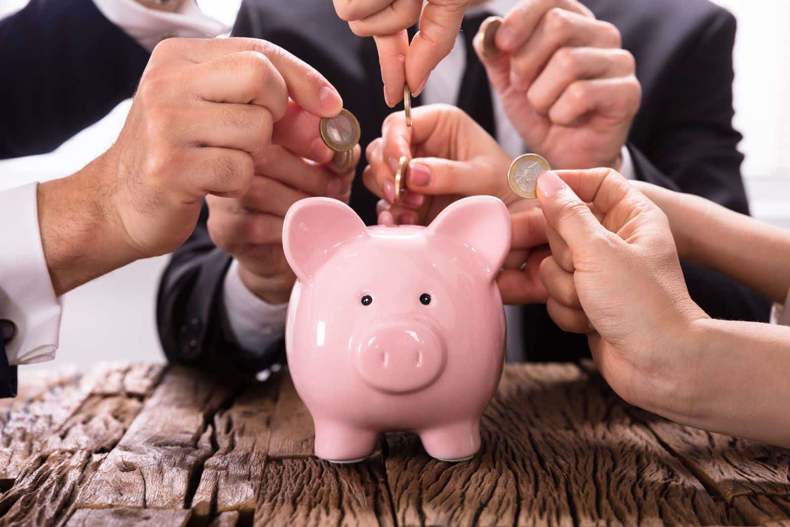 Five business people feed coins into a single piggy bank.