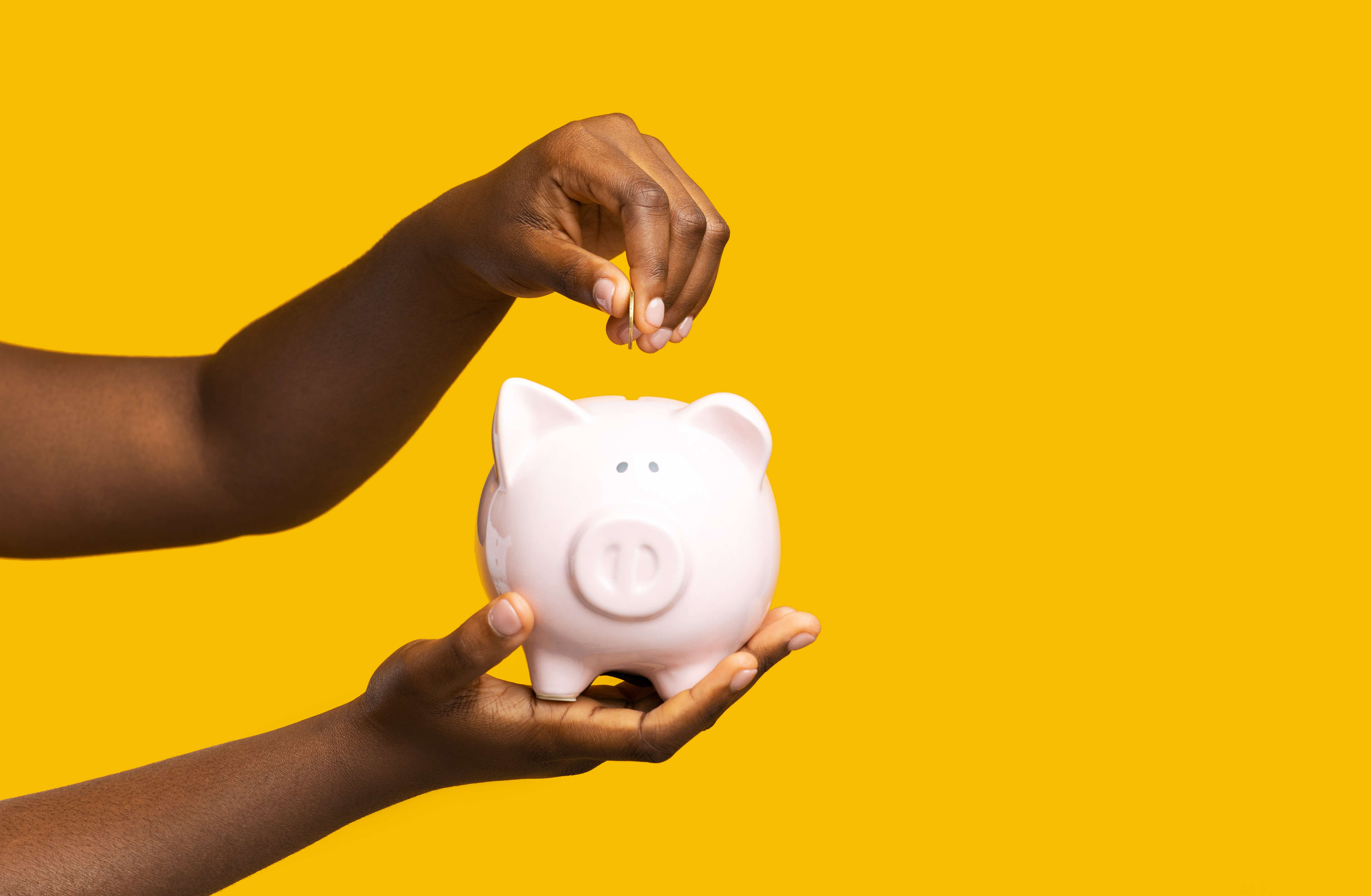 Someone putting a coin into ceramic piggy bank over yellow studio background