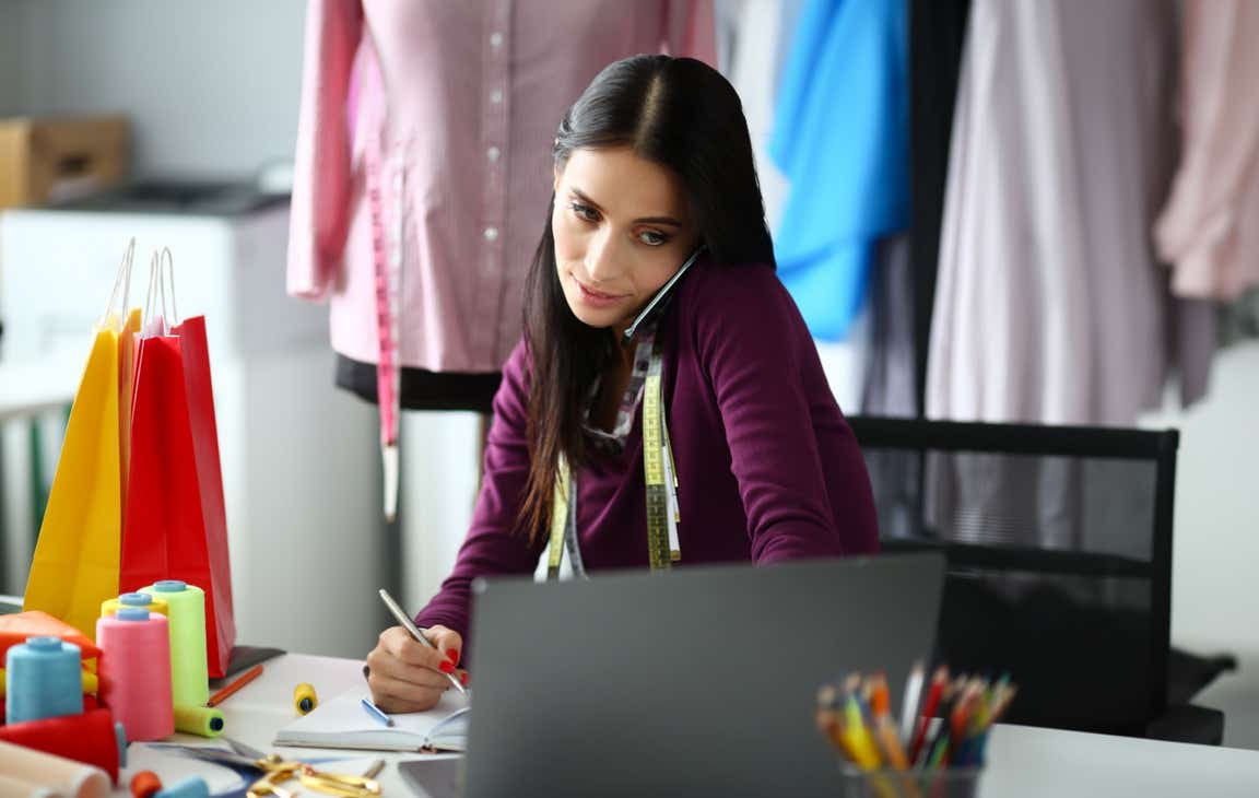 Busy clothing designer holds her phone to her ear with her shoulder while using her laptop and taking notes with a pen.