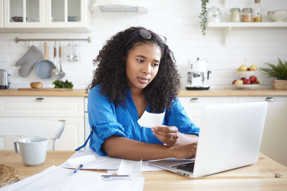 Woman is sitting at her laptop looking at the screen and holding a receipt. 