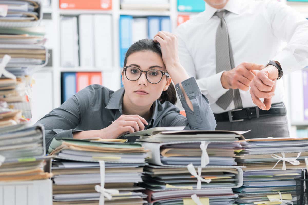 A woman in glasses sits in front of piled of work while a man taps his watch behind her