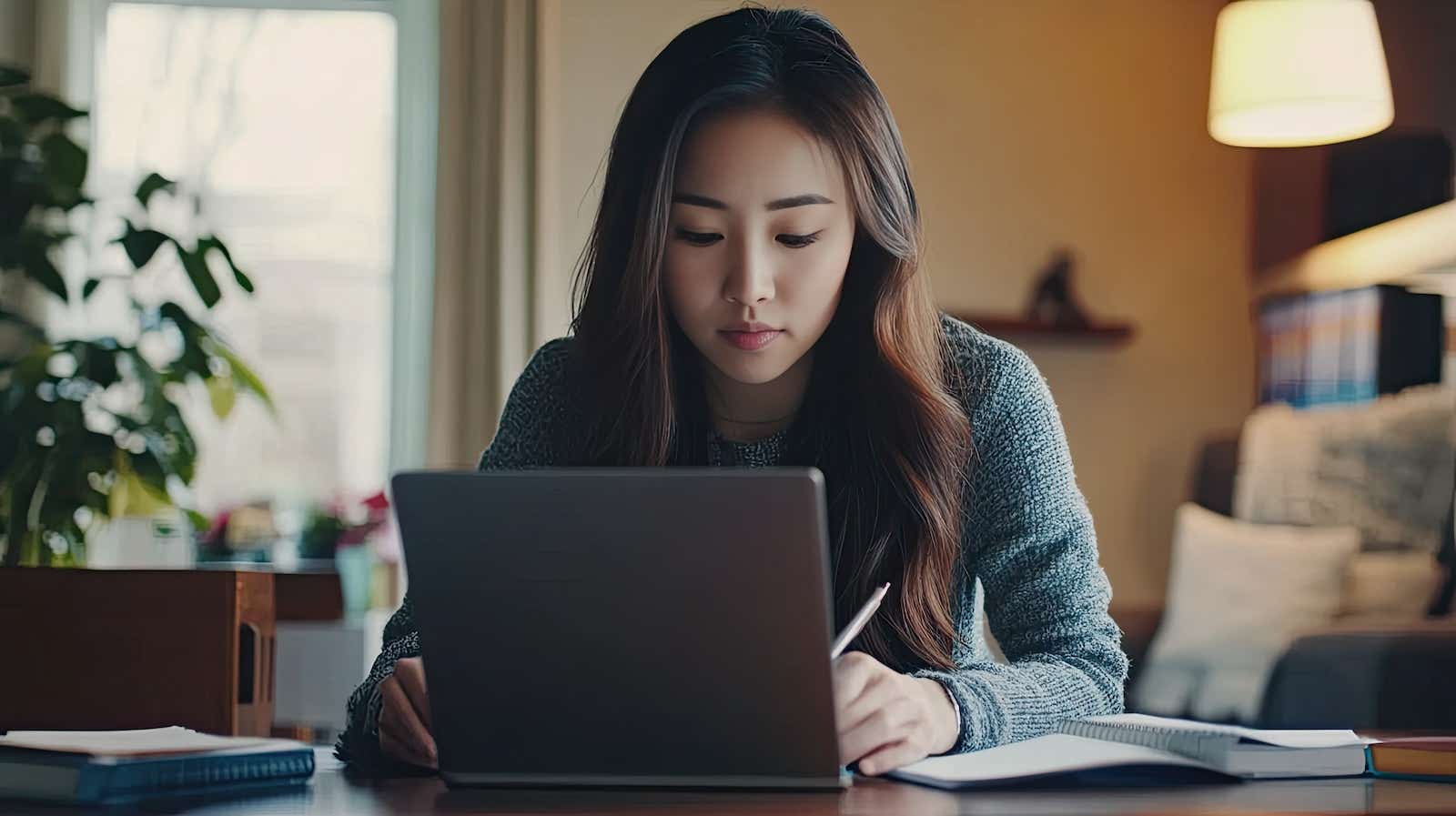 Home office environment with young Asian woman working on laptop and taking notes.