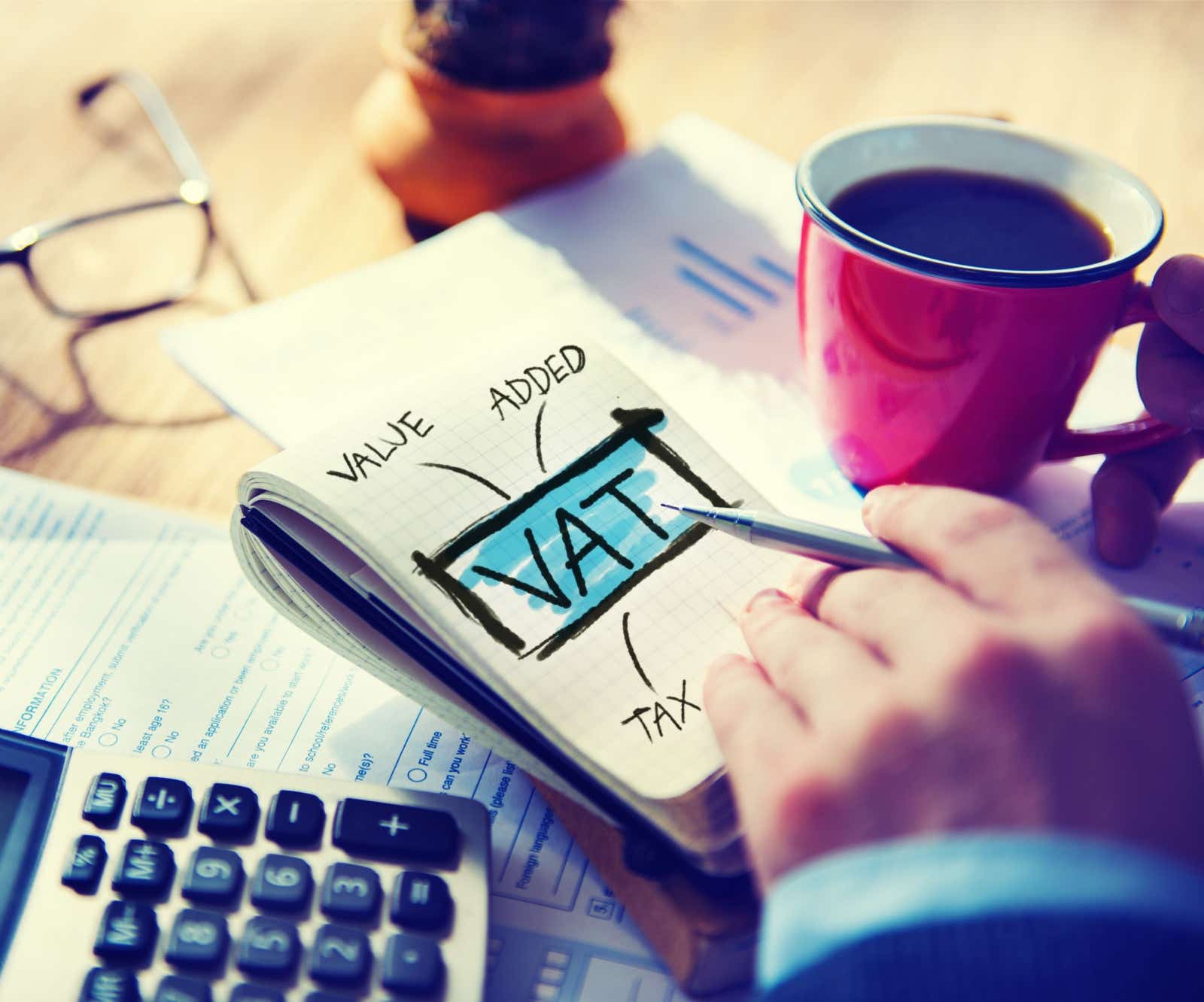 Notepad with VAT written on it. Beside it on the desk is a cup of black coffee a form and a calculator.