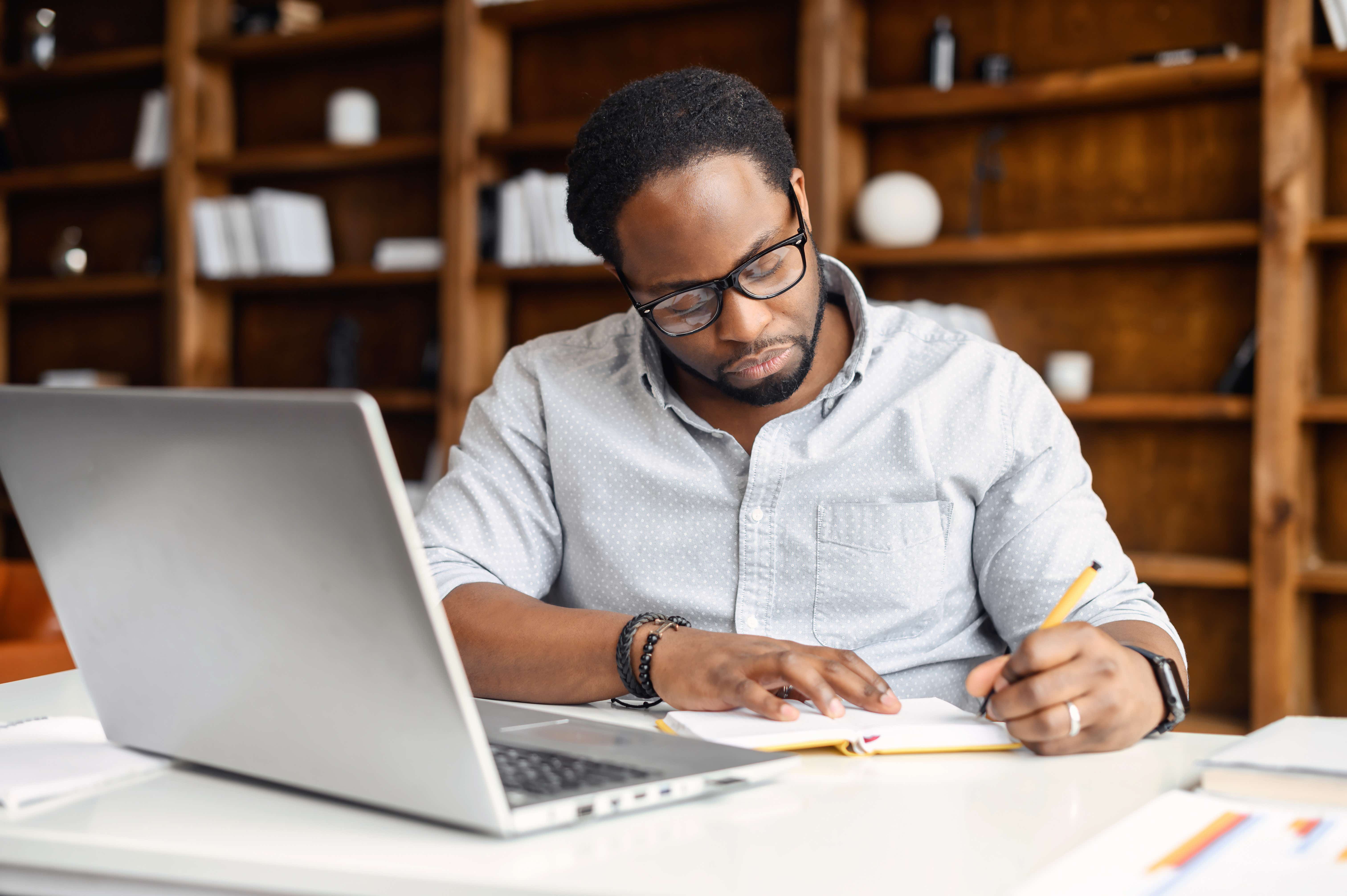 Man writes in a book. In front of him is an open laptop.