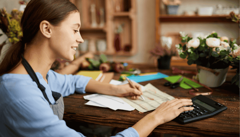 Small-business-owner-in-a-flower-shop-smiling-while-working-at-a-desk,-using-a-calculator-and-reviewing-documents