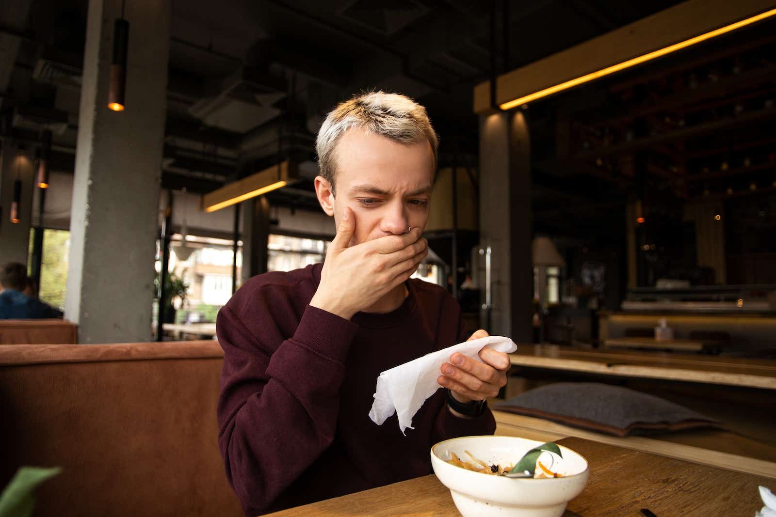 A young man in a cafe covers his mouth and looks in disgust at the food in front of him. 