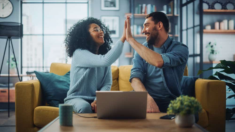 A couple are sitting on a sofa with a laptop in front of them and they are high-fiving 