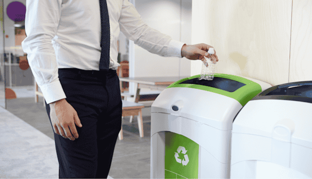 Person-in-business-attire-recycling-a-plastic-bottle-into-a-bin-labeled-'mixed-recyclables'-inside-a-workplace-environment