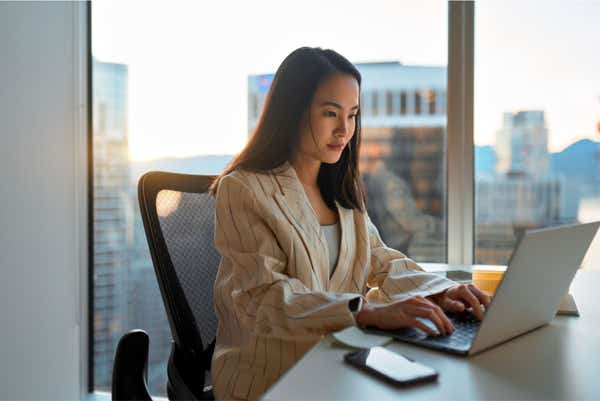 Young business woman working on her laptop. Alongside her desk is a big picture window showing a cityscape.