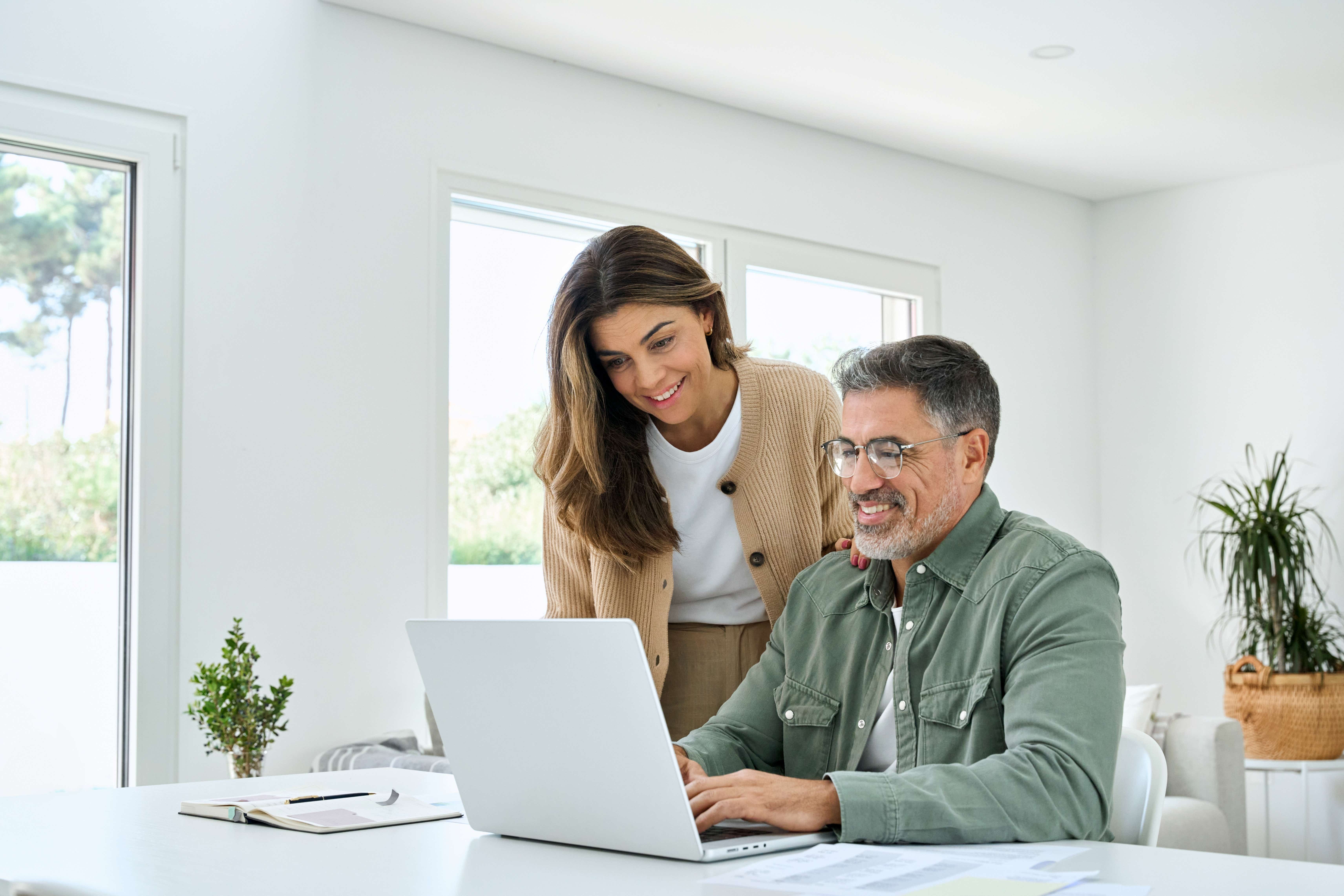 Smiling middle aged senior man working on computer sitting at table with wife standing nearby in living room. 