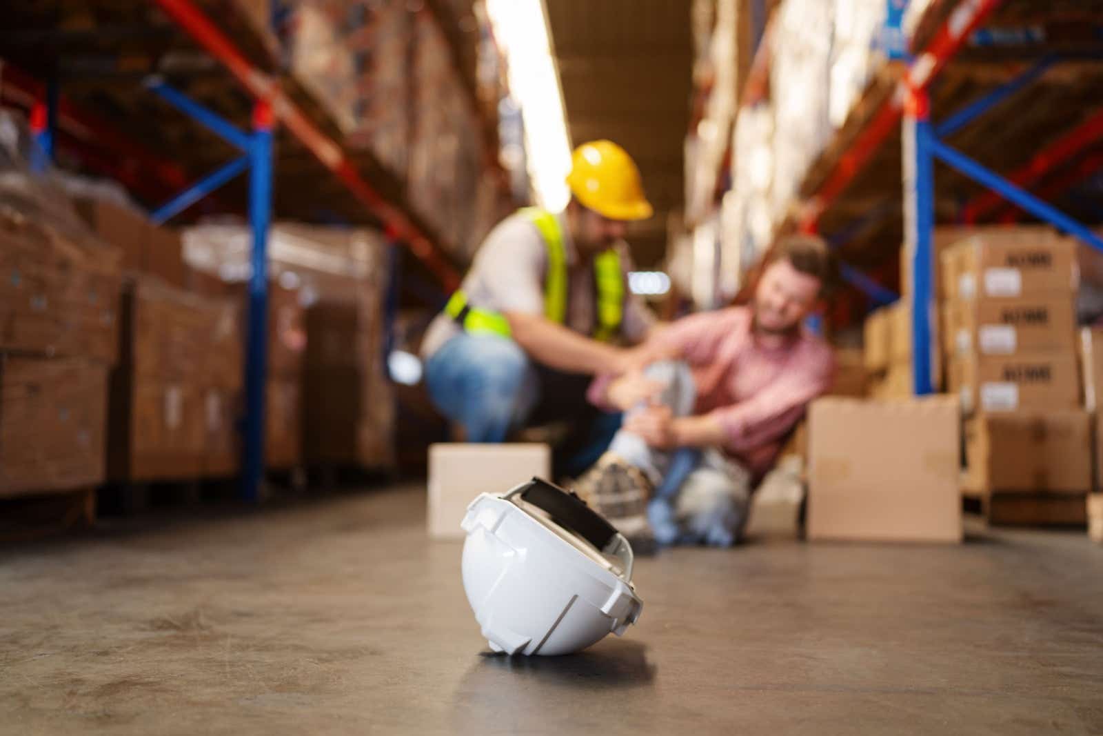 Scene in a warehouse. A worker holds his leg in pain, while a colleague tries to help him.