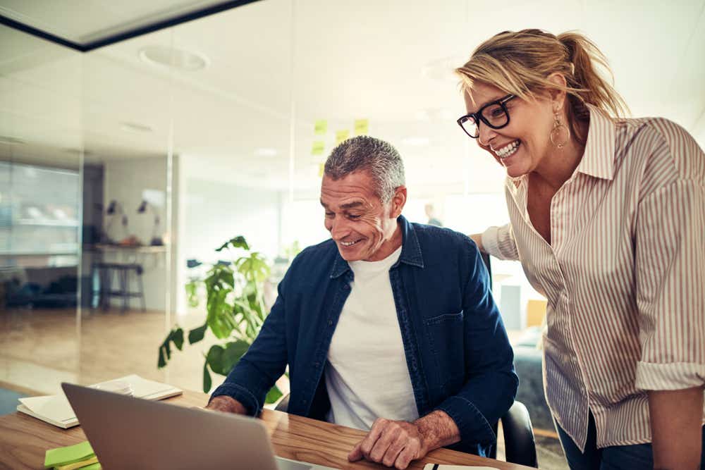 Man and woman at home smiling as they look at a laptop. Man is sitting at the desk and woman is standing next to him. 