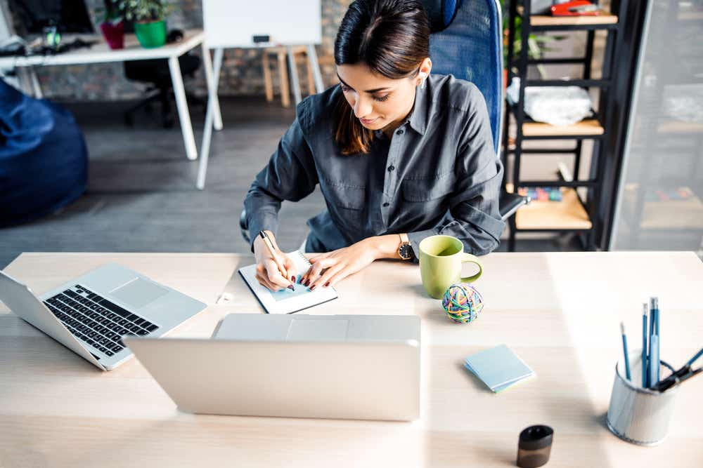 Women at desk writing on a notepad with a laptop nearby